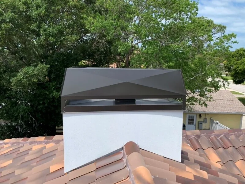 Dark metal chimney cap on stucco flue above clay tile roof